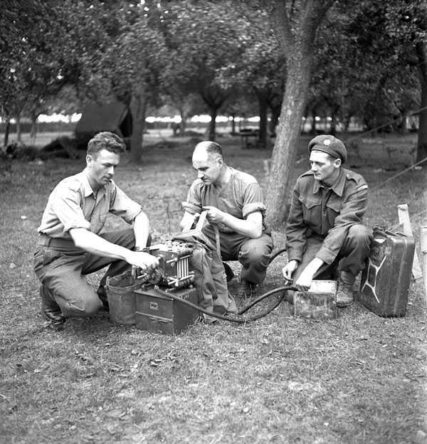 Photographie en noir et blanc – Trois soldats sont accroupis au sol, en train de travailler à une petite pièce de machinerie.