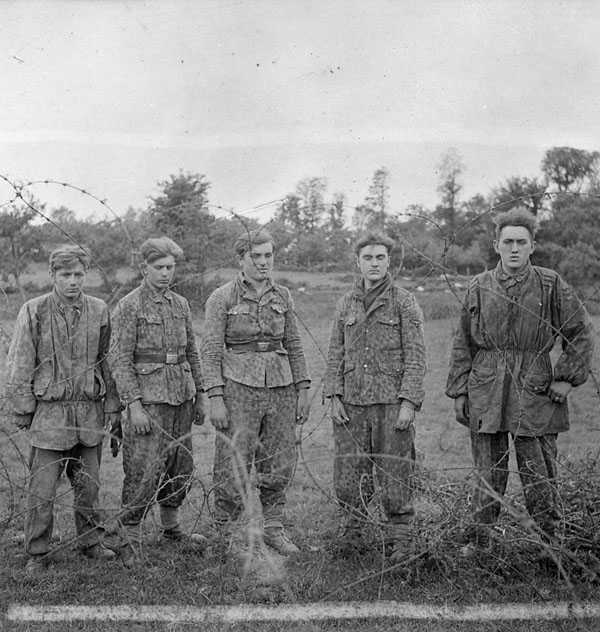 Photographie en noir et blanc – Cinq très jeunes soldats allemands en diverses tenues militaires se trouvent derrière une épaisse clôture de fil barbelé.