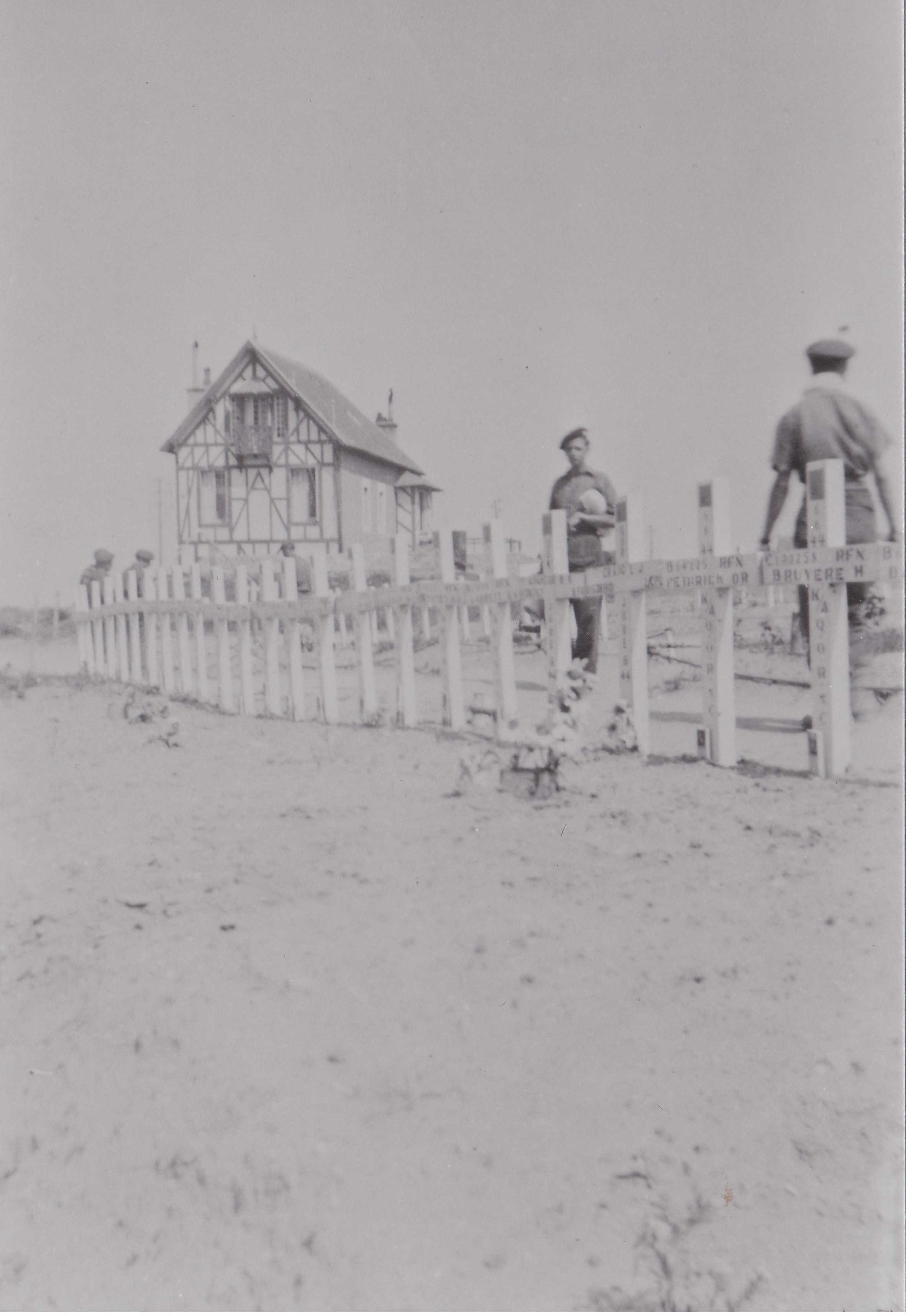 Photographie en noir et blanc – Une maison typiquement française est visible à l’arrière-plan. Deux hommes marchent derrière une série de croix blanches érigées sur le sol. La photo est délavée, mais on voit que des noms sont inscrits sur chaque croix.