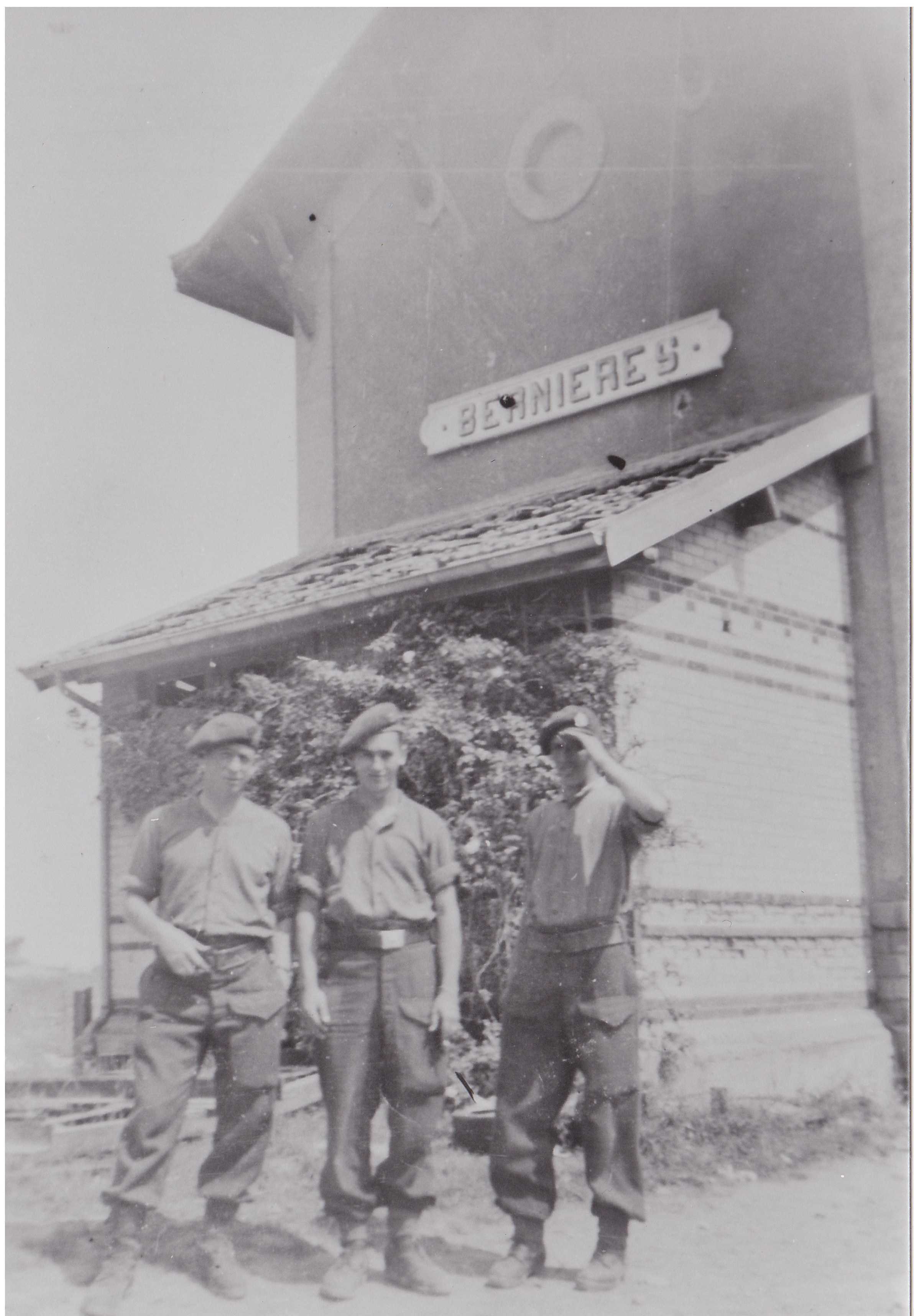Photographie en noir et blanc – Trois Canadiens dans une tenue militaire décontractée, debout en face d’un grand bâtiment indiquant « Bernières ». L’un a abaissé sa casquette pour se protéger contre le soleil.