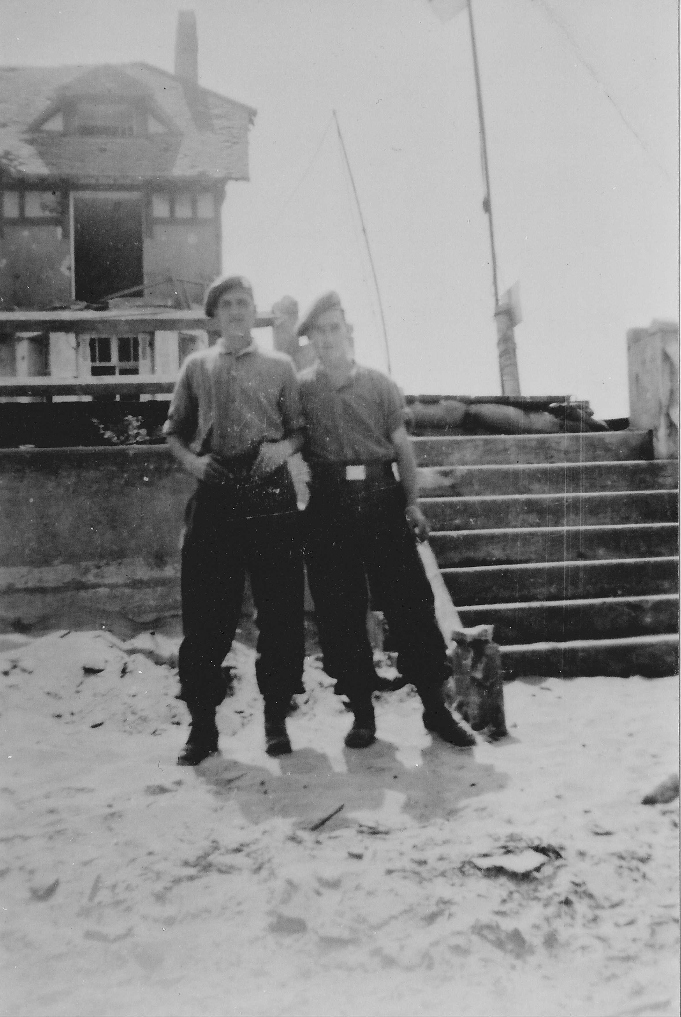 Photographie en noir et blanc – Deux soldats canadiens debout devant la digue; un escalier en pierre est à leur gauche. Une maison est visible à l’arrière-plan; l’une des fenêtres a explosé. Ils posent debout, sur une plage sablonneuse.