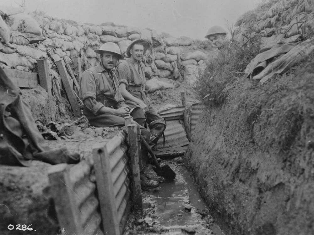Photographie en noir et blanc – Deux soldats canadiens sont assis sur le rebord d’une tranchée boueuse, alors qu’un troisième se trouve derrière un coin. Le fond de la tranchée est couvert d’eau.