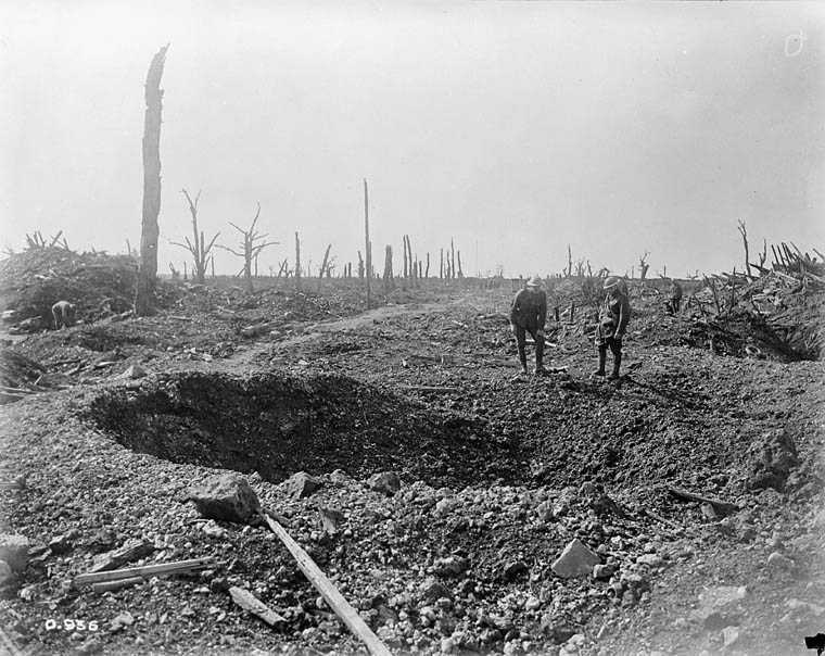 Photographie en noir et blanc – Un grand trou d’obus perce le milieu d’un chemin. Deux hommes en uniforme militaire et portant des casques se tiennent derrière, l’un étant légèrement accroupi. Le paysage est rude et rocheux.