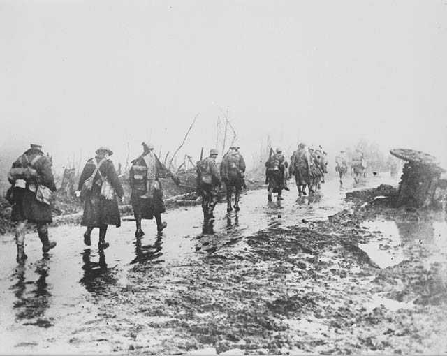 Photographie en noir et blanc – Un groupe de soldats s’éloigne de la caméra en file indienne. La route est couverte de boue et d’eau. Les soldats portent des paquets sur leur dos.