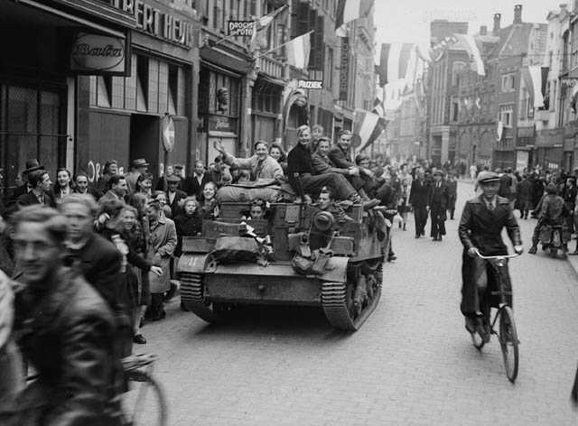 Photographie en noir et blanc – Scène de rue. Des drapeaux néerlandais sont partout, flottant ou brandis. La foule se masse dans les rues et les trottoirs, à pied et en vélo. Un char roule au centre, et des civils y sont montés.