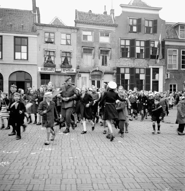 Photographie en noir et blanc – Place publique fourmillant de monde, bâtiment étroit et un drapeau à l’arrière-plan. De nombreux enfants en manteaux circulent un peu partout sur la grande place, enthousiastes. Bon nombre agitent un drapeau hollandais.