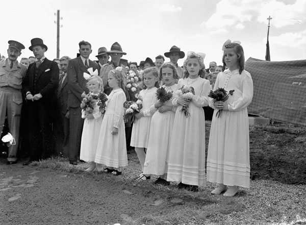 Photographie en noir et blanc – Six jeunes filles néerlandaises sont debout, en ligne. Elles portent des robes blanches et tiennent des bouquets de fleurs. Des passants en tenue civile ou militaire se trouvent derrière elles.
