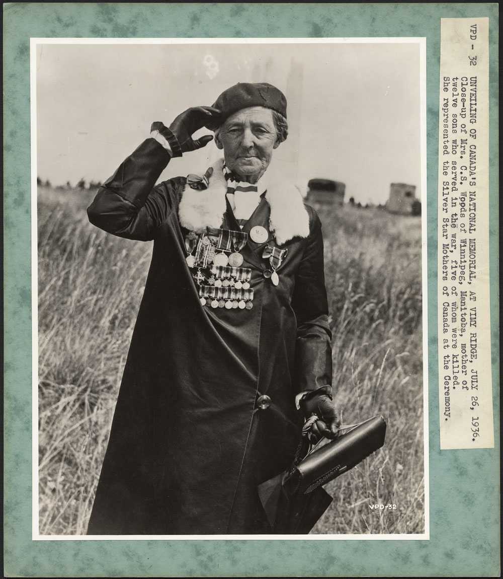 Photographie en noir et blanc, bordée de vert. Une femme âgée portant un béret salue la caméra de la main droite. Elle porte un long manteau noir garni de fourrure et orné de médailles, dont certaines sont miniatures.