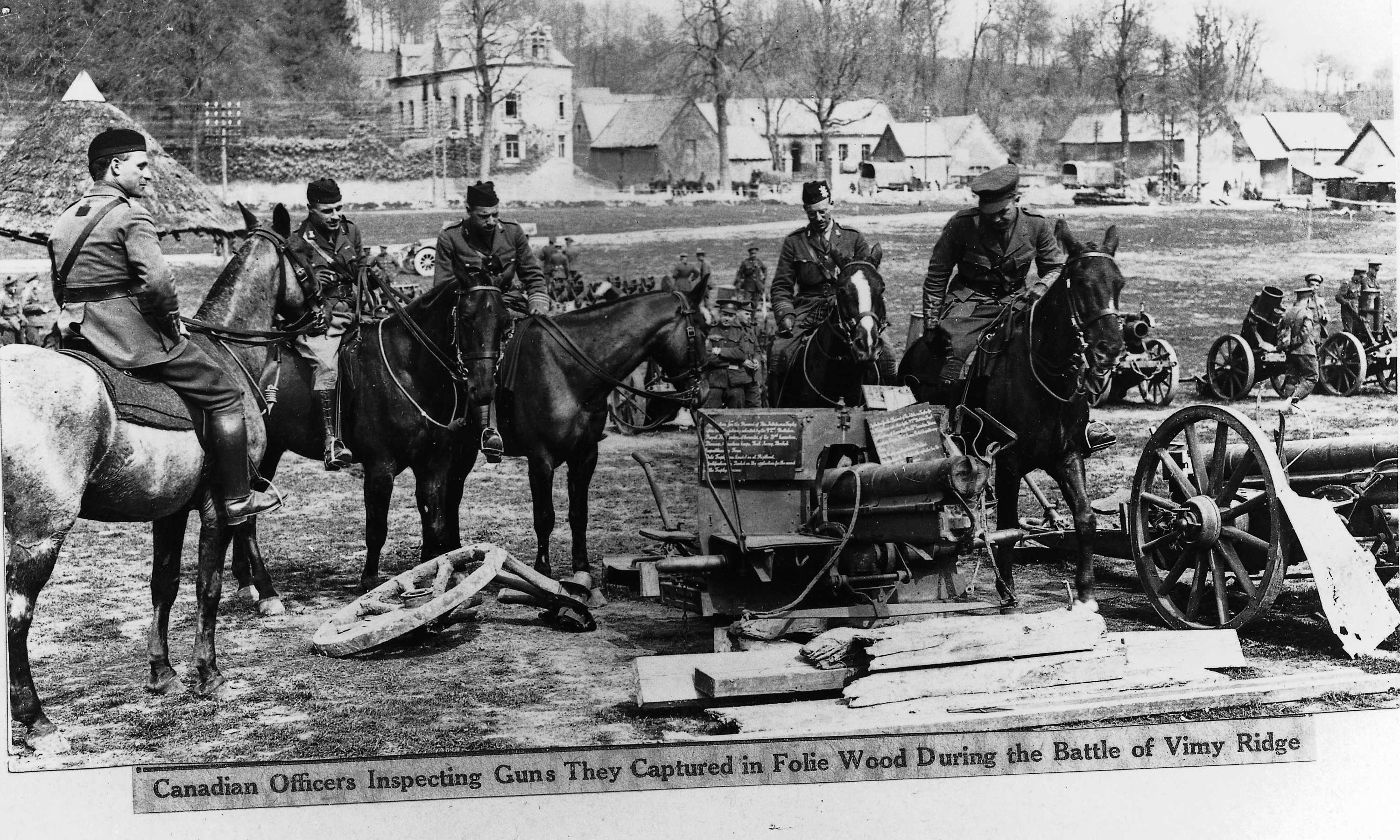 Photographie en noir et blanc – Du haut de leurs chevaux, des hommes en uniforme militaire regardent une pile de canons montés sur roues pour en faciliter le déplacement. De gros morceaux de bois gisent sur le sol. Un village est visible au loin.