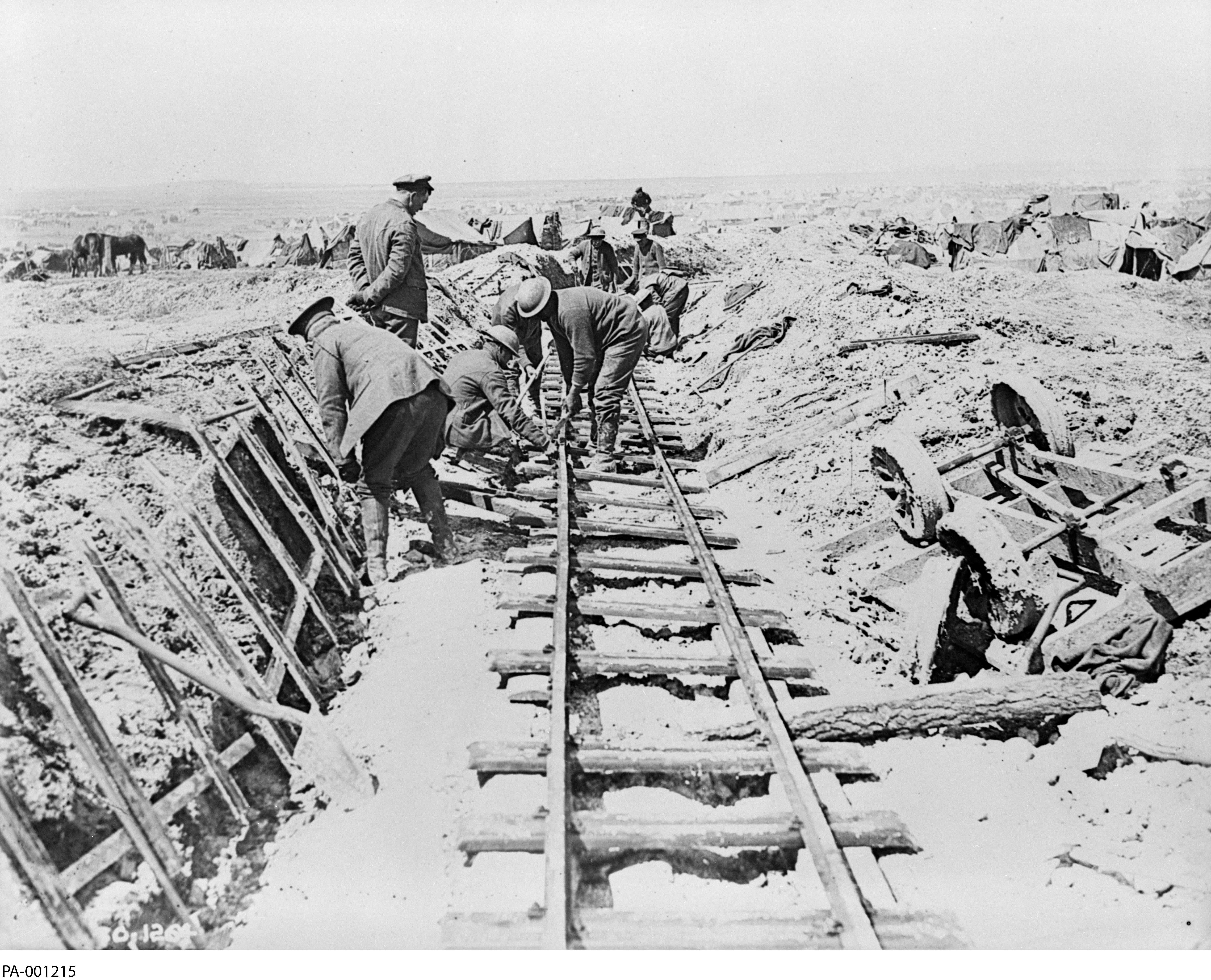 Photographie en noir et blanc - À l'avant-plan, sur un sol boueux, quatre hommes posent des rails pré-assemblés. Un cinquième homme, en tenue civile, les observe.